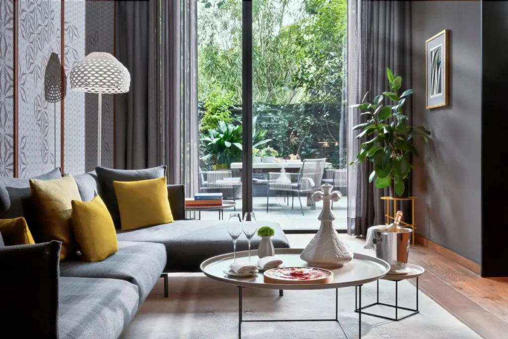 Gray sofa with yellow cushions and a coffee table beside glass doors opening to a planted terrace in a Terrace Suite at Mandarin Oriental, Barcelona.