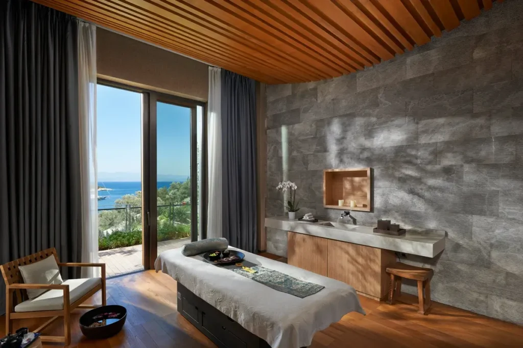 Treatment bed, stone walls, timber ceiling, and a tall window facing the sea in a spa treatment room.