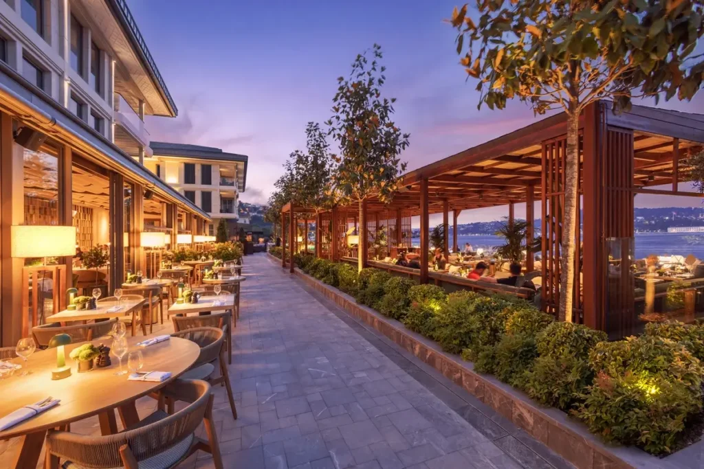 Outdoor tables beside the hotel and illuminated waterfront planting at Olea & Bar, Mandarin Oriental Bosphorus, Istanbul.