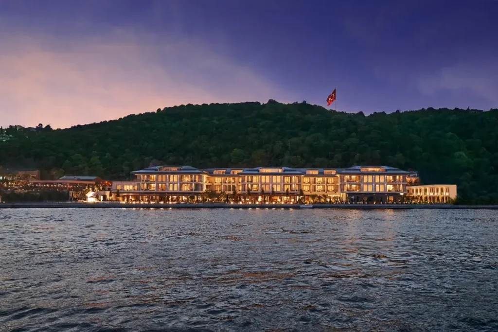Illuminated buildings of Mandarin Oriental Bosphorus, Istanbul beside the strait, with a wooded hillside behind them at dusk.