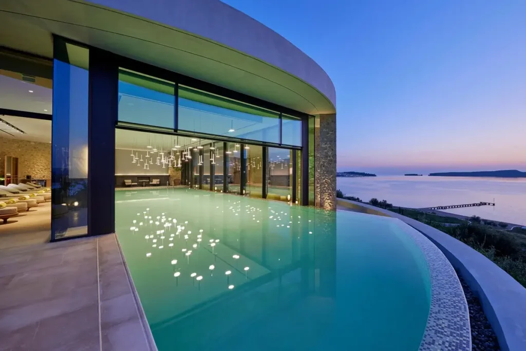 Curved spa pool extending from a glass-fronted building toward Navarino Bay at dusk.