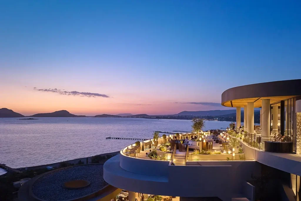 Illuminated outdoor tables on the curved Three Admirals Lounge terrace overlooking the bay at dusk.