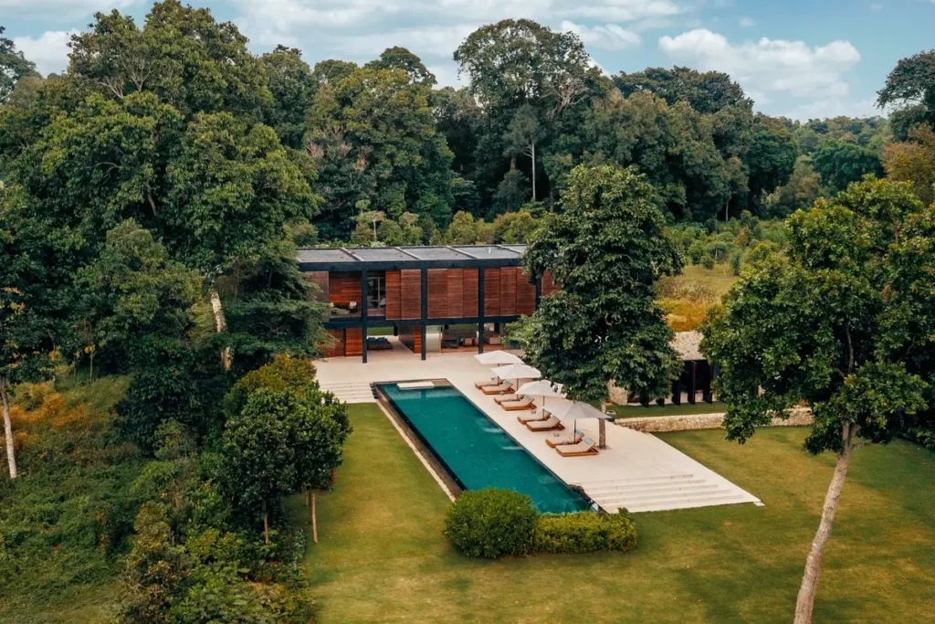 Aerial view of a standalone villa, long private pool, loungers, and lawn enclosed by coastal forest.