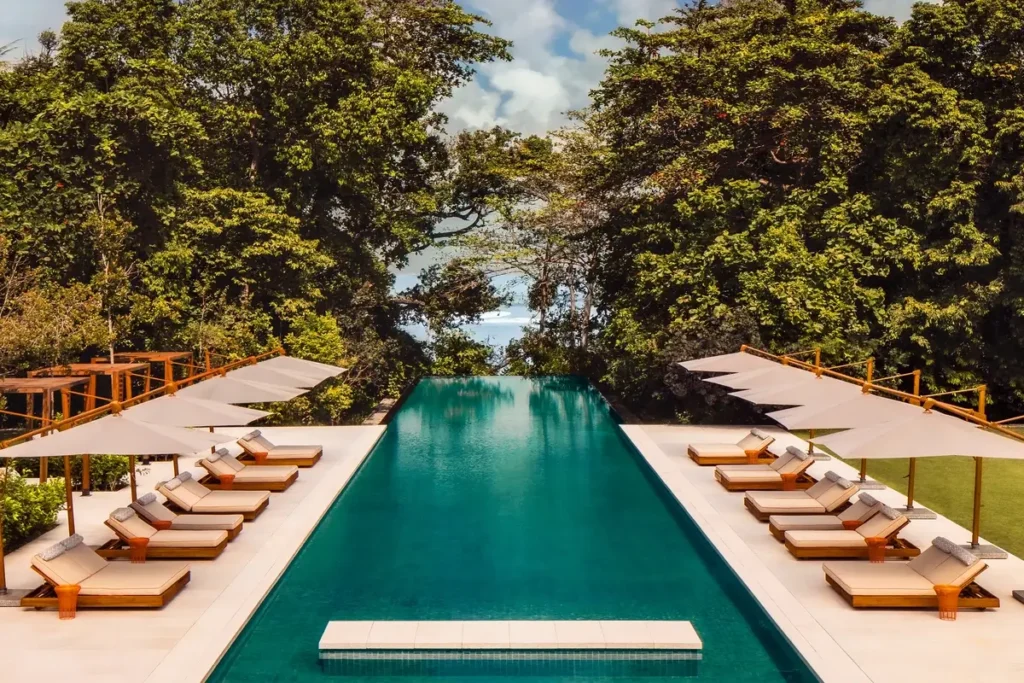 Long swimming pool lined with loungers and umbrellas, surrounded by dense trees at Mandarin Oriental, Desaru Coast.