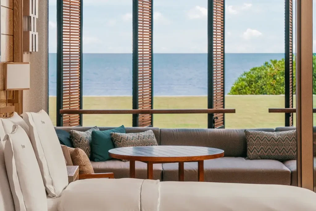 Sofa and round table beside tall windows overlooking a lawn and the sea in an Ocean Front Junior Suite.
