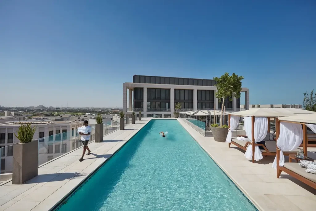 Long rooftop swimming pool with a glass balustrade, white cabanas, and city buildings beyond.