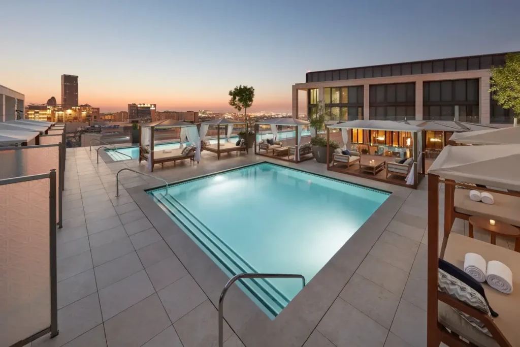 Rectangular rooftop pool surrounded by loungers and cabanas beneath a sunset sky.