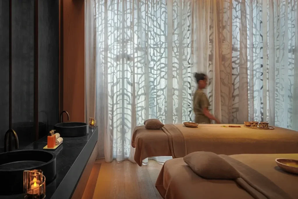 Two treatment beds, patterned curtains, and a therapist in a softly lit spa room.