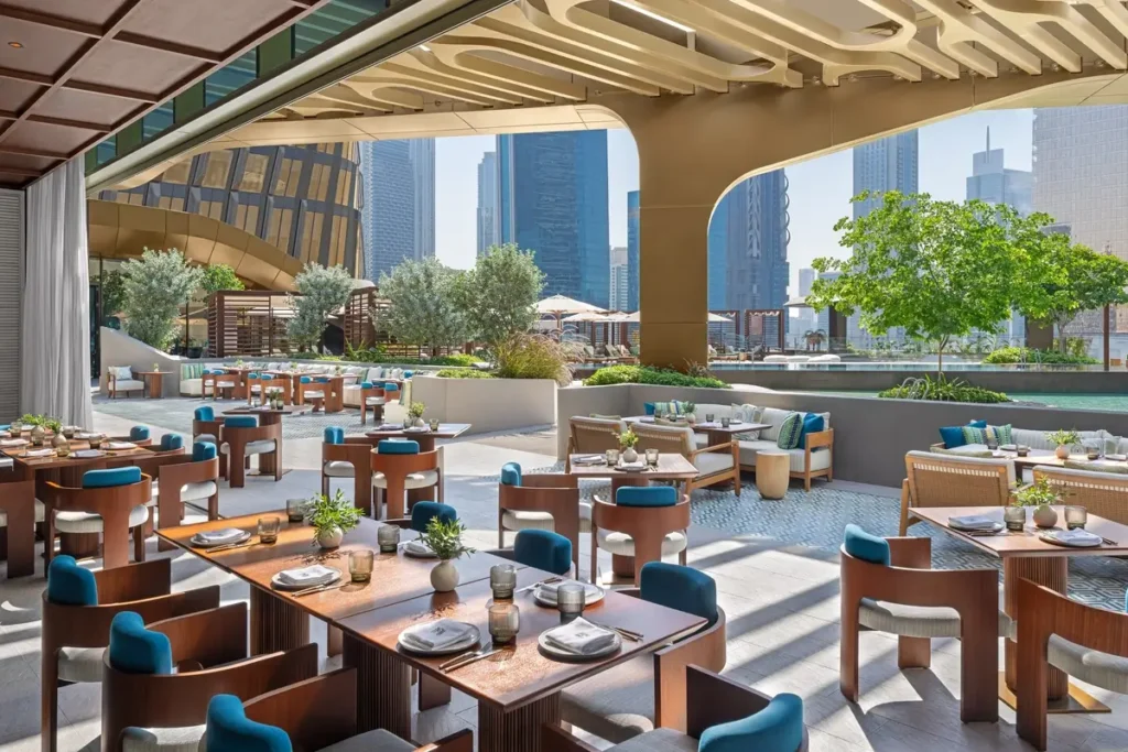 Set tables, blue-accented chairs, trees, and shade structures on Noia’s outdoor terrace.