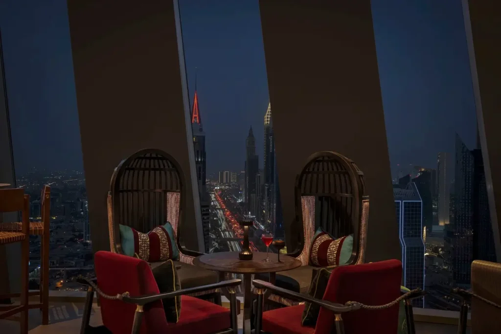 Red chairs, hanging seats, and tables beside tall windows overlooking Dubai’s illuminated skyline at Yù & Mì.