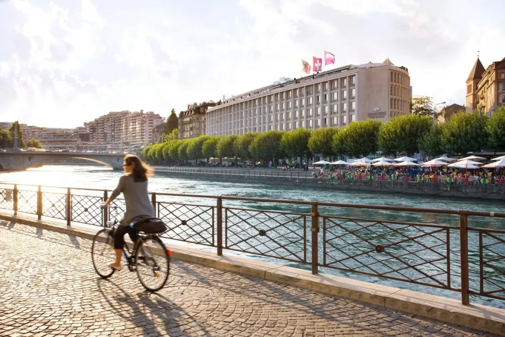 Cyclist on a riverside path opposite Mandarin Oriental, Geneva, with the Rhône and hotel facade behind.