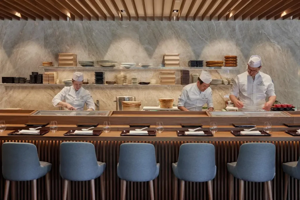 Chefs preparing food behind SACHI’s wooden omakase counter, with upholstered seats in front.
