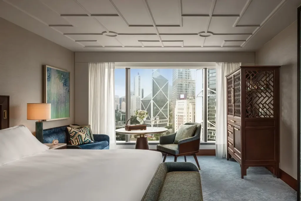 Bed, sitting area, decorative screen, and windows toward Central’s towers in a Statue Square View Room.