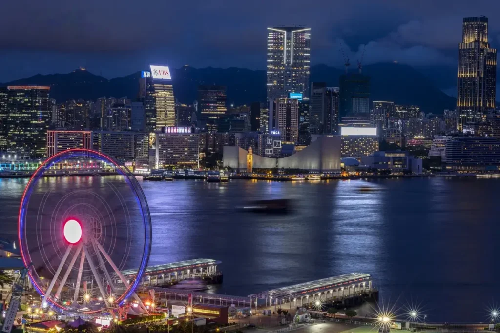 Victoria Harbour at night, with the Hong Kong Observation Wheel in the foreground and illuminated buildings across the water.