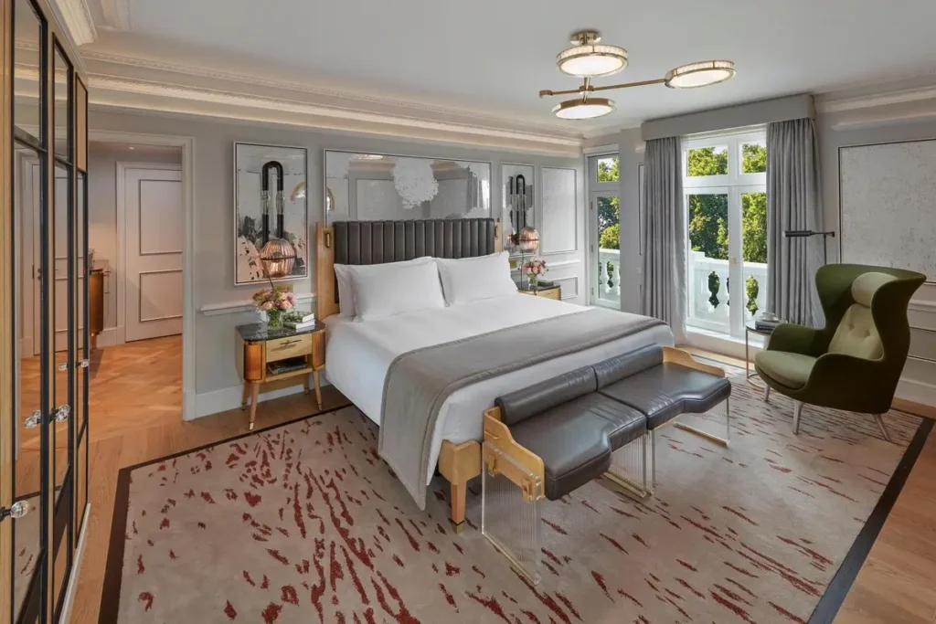 Bed, upholstered bench, armchair, and tall windows facing trees in a Hyde Park Room.
