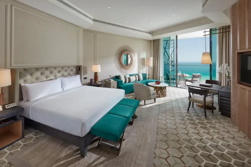 Bed, turquoise seating, dining table, and balcony doors facing the Gulf in a Deluxe Sea View Room.