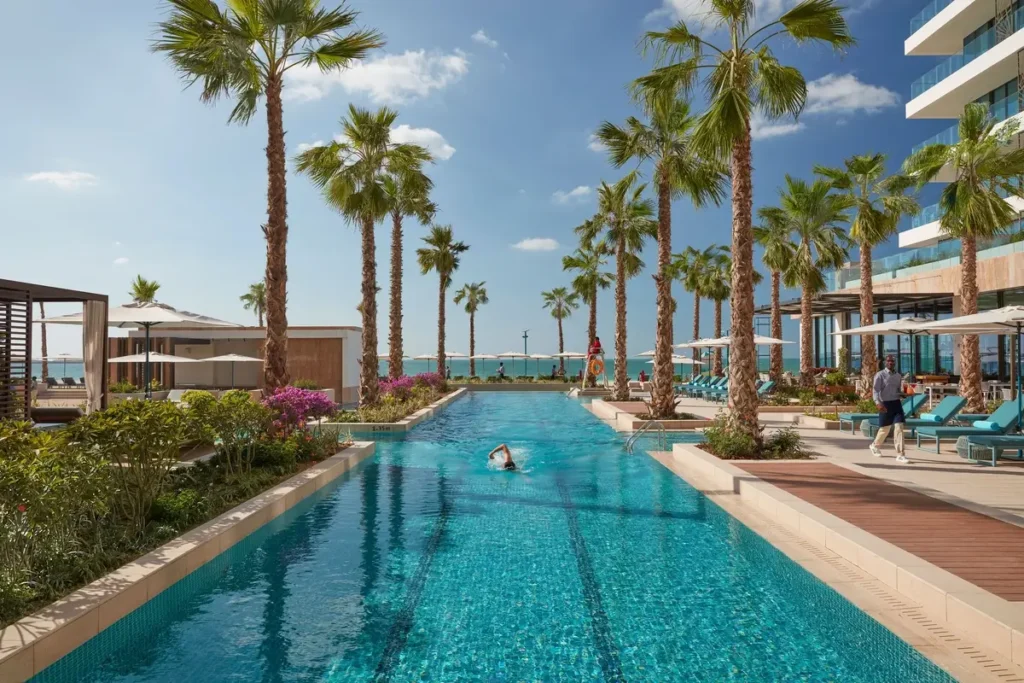 Long outdoor pool lined with palms, loungers, and resort terraces, with the sea beyond.