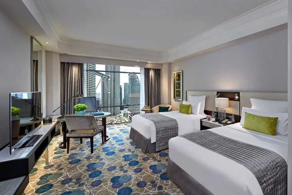 Twin beds, a desk, and seating beside windows facing the Petronas Towers in a Deluxe Twin Towers View Room.