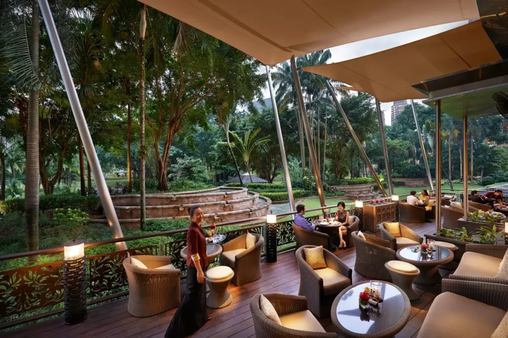 Outdoor lounge chairs and tables beneath awnings beside lush park planting.