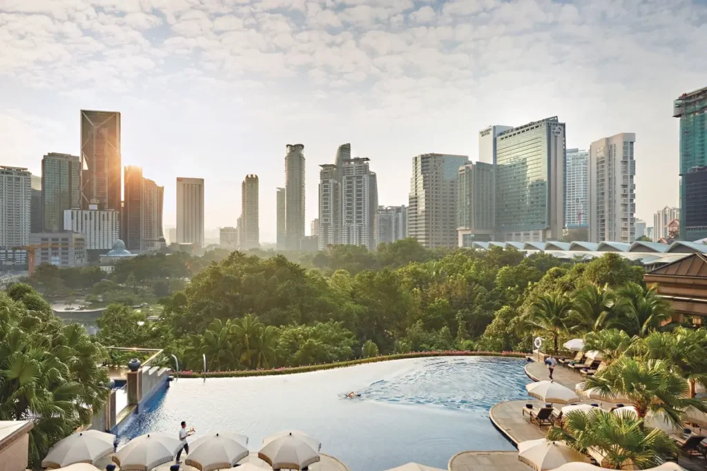 Curved outdoor pool with loungers, palms, KLCC Park greenery, and city towers beyond.
