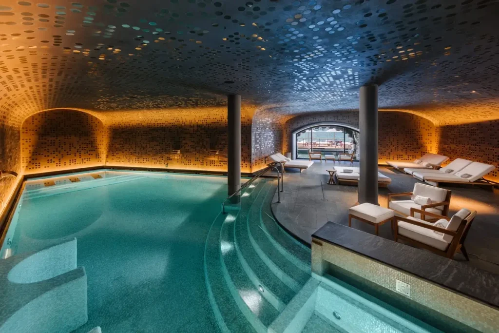 Curved indoor pool with hydrotherapy areas, loungers, and warm lighting beneath a reflective ceiling.