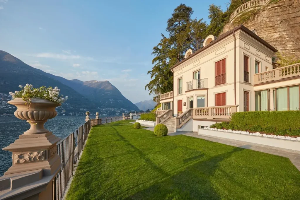 Pale villa facade with red shutters beside a lawn and stone balustrade overlooking Lake Como.