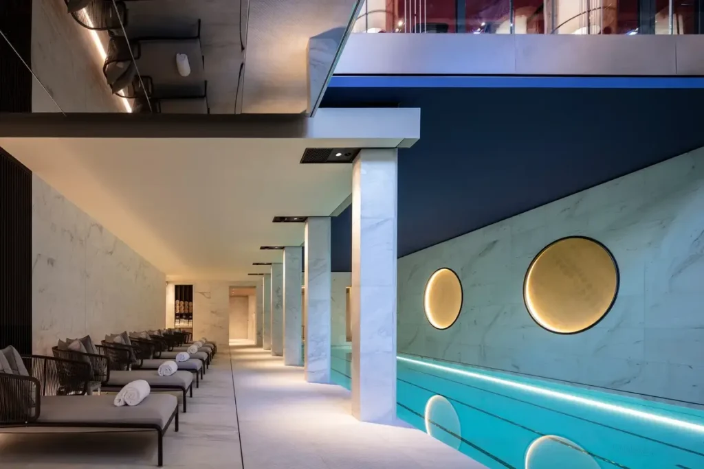 Indoor pool with illuminated blue water, circular wall details, pale columns, and a row of loungers.