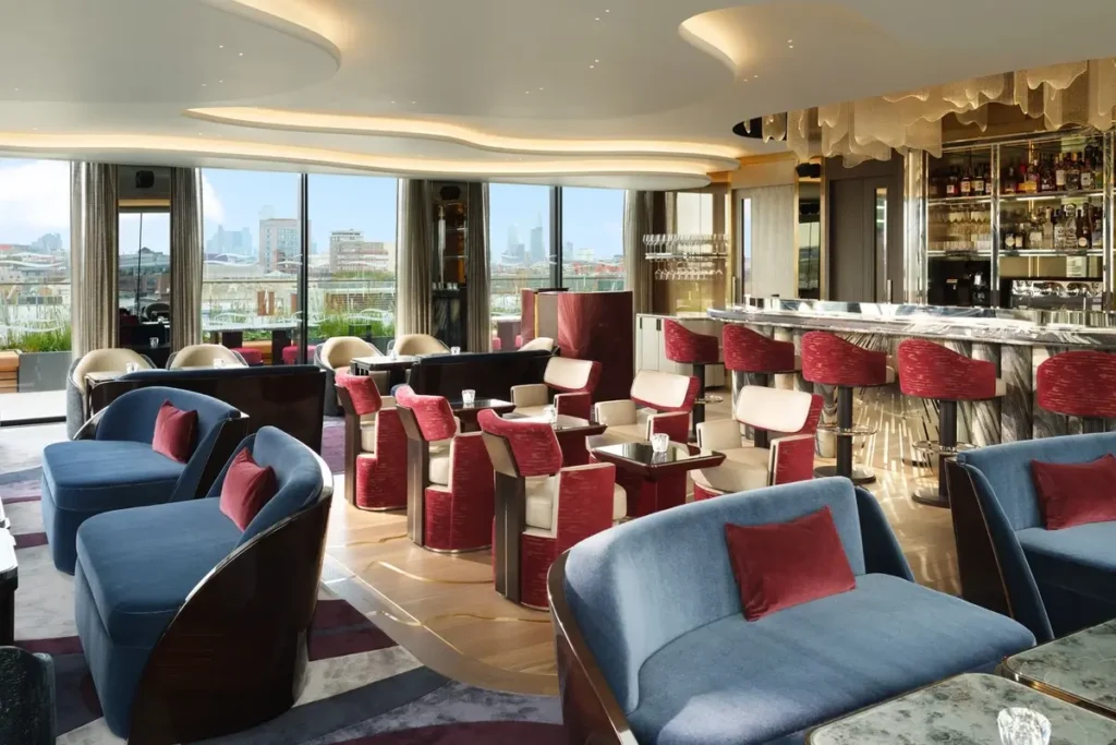 Blue armchairs, red bar stools, and a gold-toned bar beside tall windows at Hanover Bar.