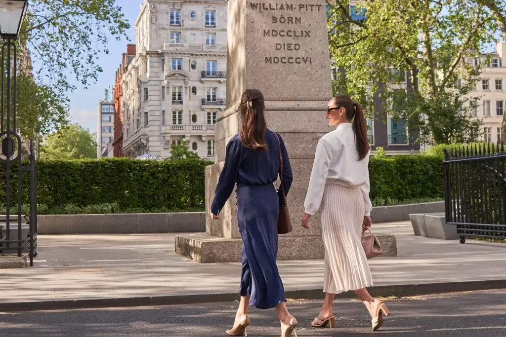Two pedestrians passing the monument and railings in Hanover Square, with trees and London buildings beyond.