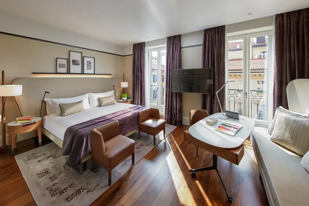 Bed, upholstered seating, and a table beside tall windows in a Deluxe Room at Mandarin Oriental, Milan.