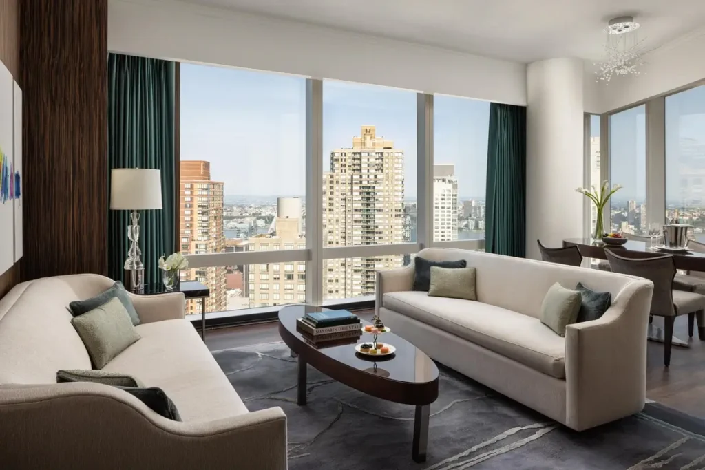 Sofas and an oval coffee table beside corner windows overlooking Manhattan in a Hudson River View Suite.