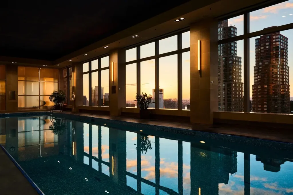 Indoor swimming pool with dark water reflecting tall windows and the Manhattan skyline at sunset.