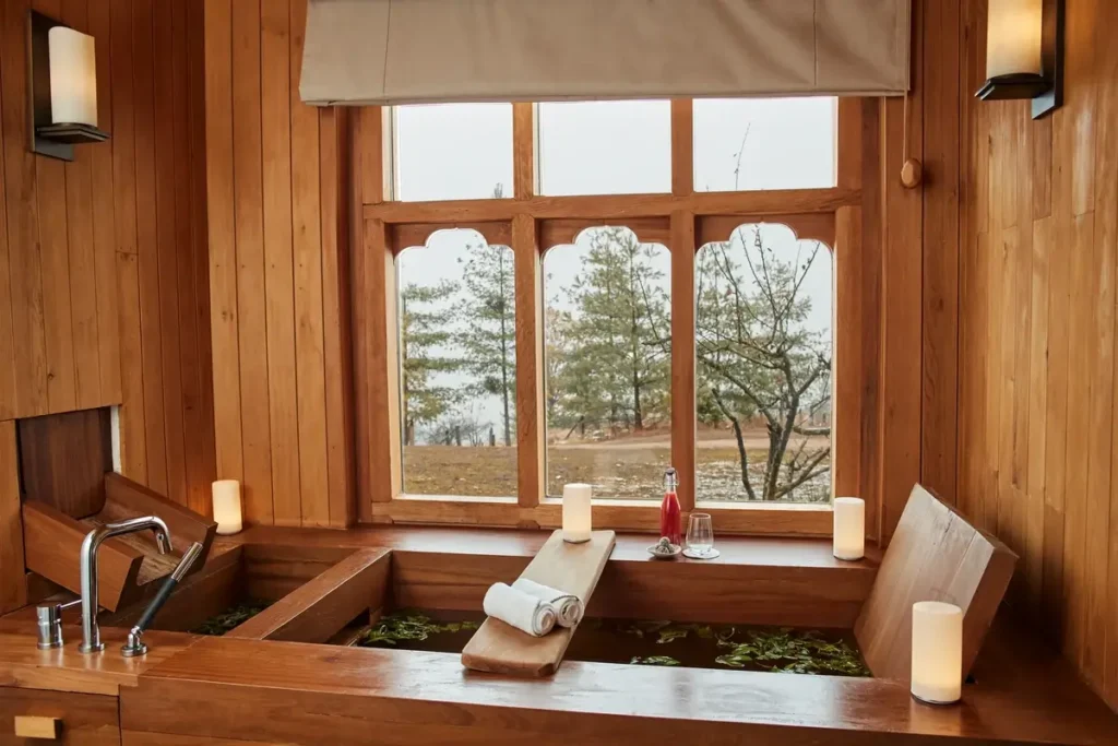 A wooden bath with a white headrest sits beneath a window overlooking trees in a softly lit timber room.