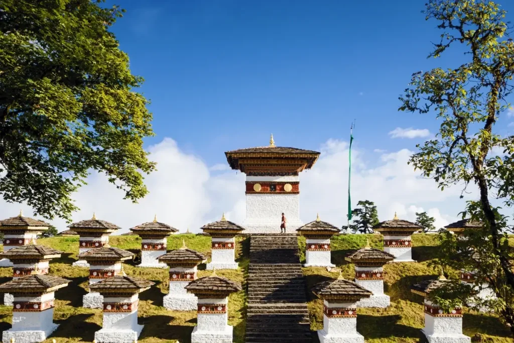 Rows of white chortens with dark tiered roofs stand beneath trees and a clear blue sky.