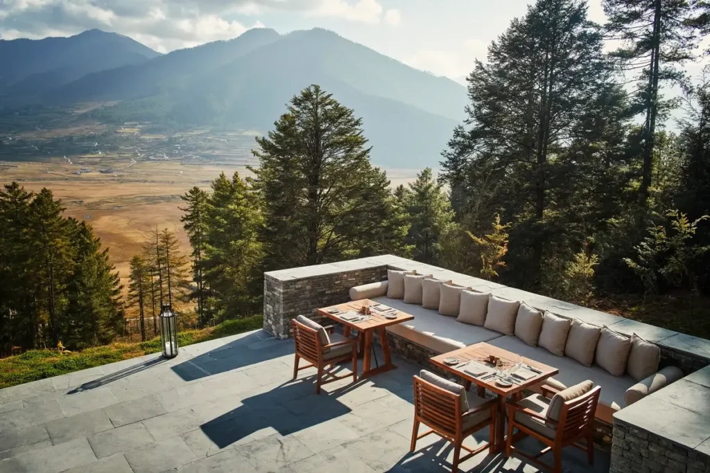 Set wooden tables and a long cushioned bench occupy a stone terrace overlooking pines and a broad mountain valley.