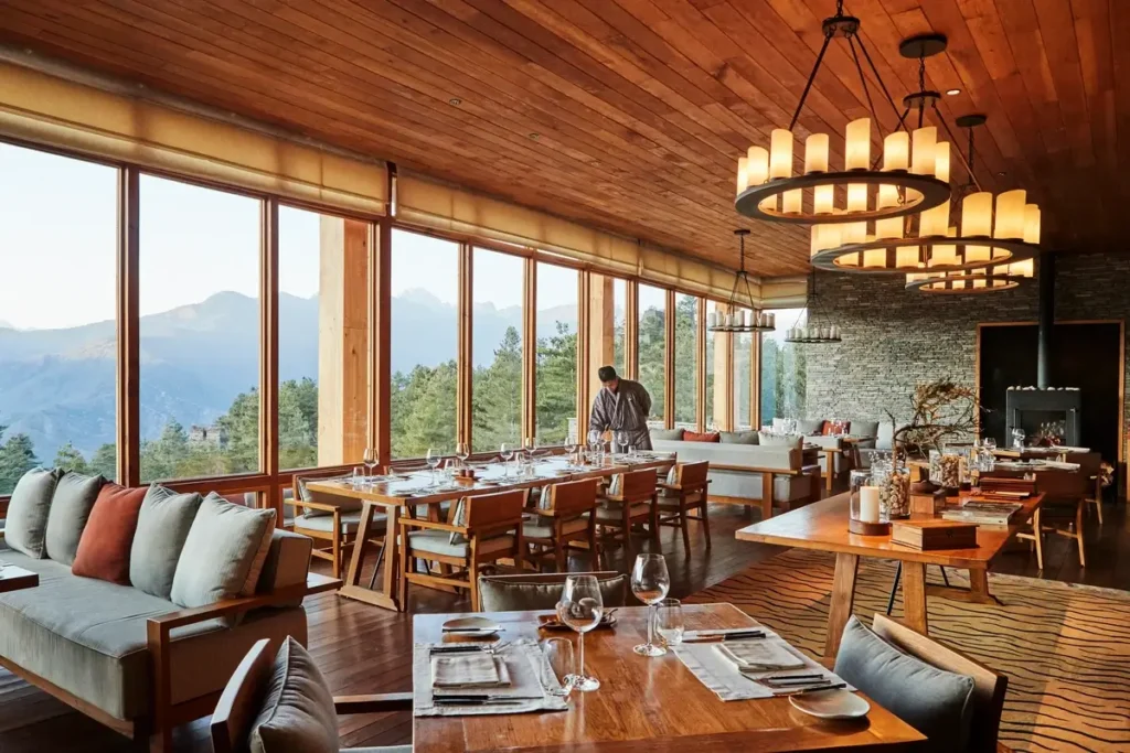 Set dining tables and cushioned seating sit beneath a timber ceiling beside large mountain-facing windows.