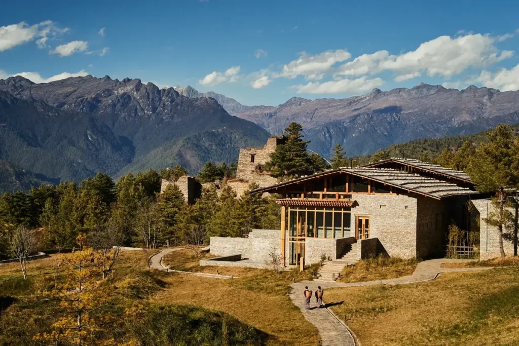 A stone lodge building stands beside old masonry ruins beneath forested ridges and high mountain peaks.