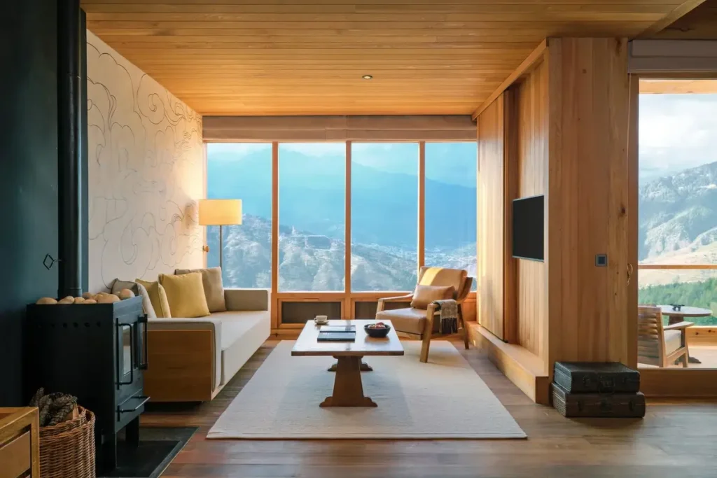 A timber-lined living room with a sofa, wood stove, and low table faces mountains through broad windows.