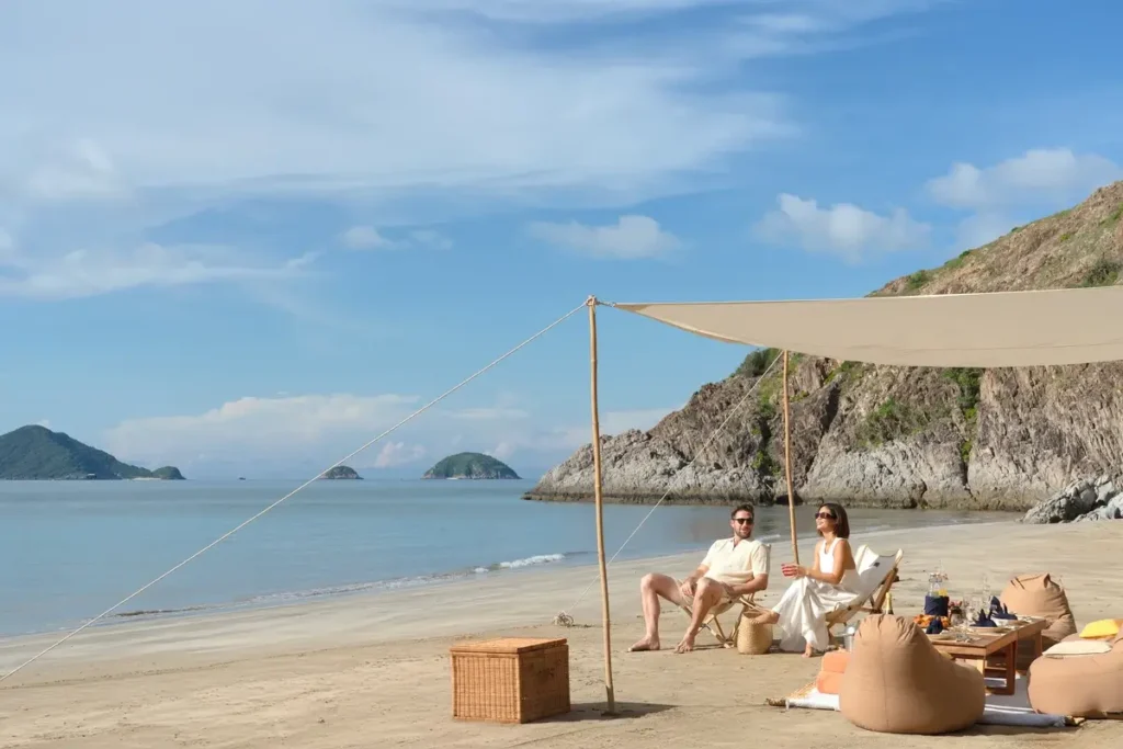 Two guests relax beneath a pale shade sail on the sand beside calm water and a rocky headland.