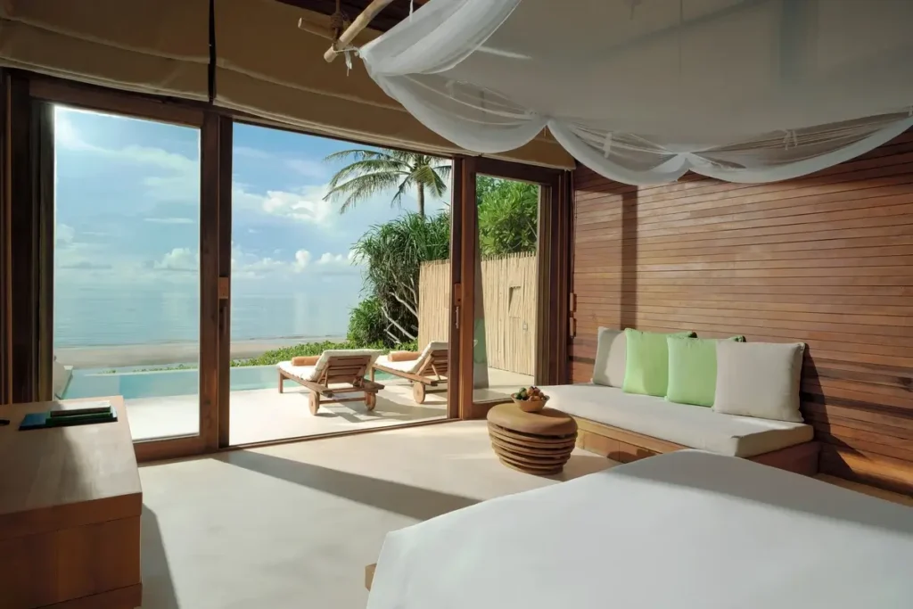 A draped bed and pale sofa sit in a timber-paneled bedroom opening toward a sunny terrace and the sea.