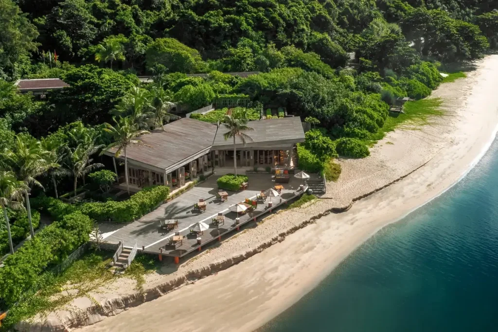 A timber-roofed restaurant and dining terrace sit beside a sandy beach backed by dense tropical trees.