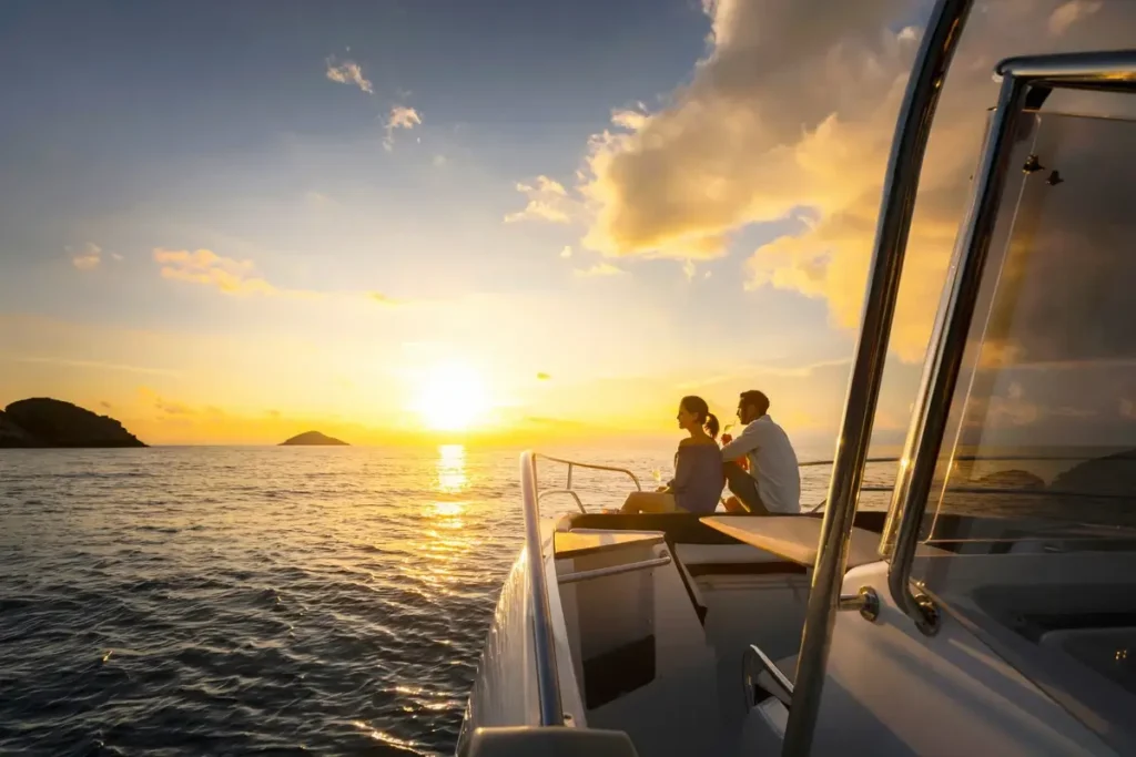 Two passengers sit at the front of a boat facing a golden sunset over the sea and distant islands.