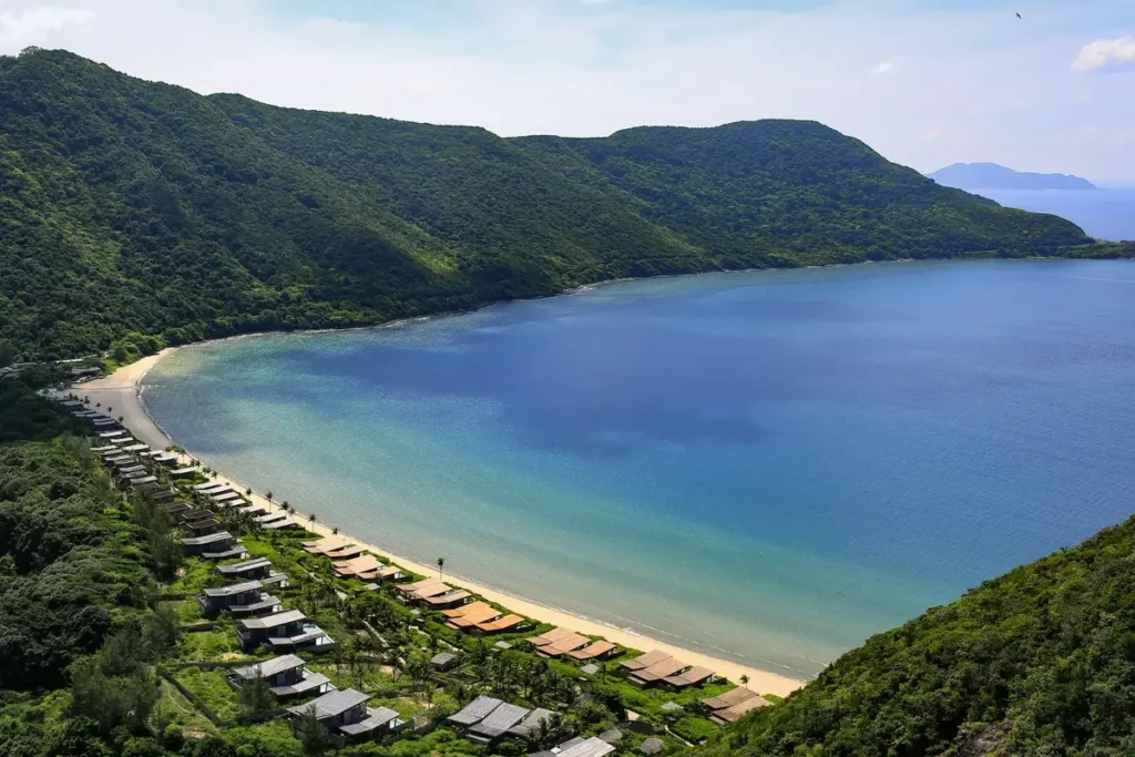 A long sandy beach curves around a blue bay beneath forested mountains, with resort villas along the shore.