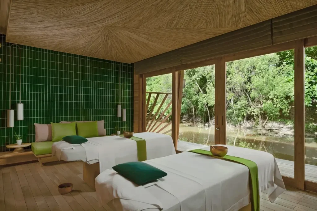 Two treatment beds with green accents face glass doors overlooking water and trees in a timber spa room.