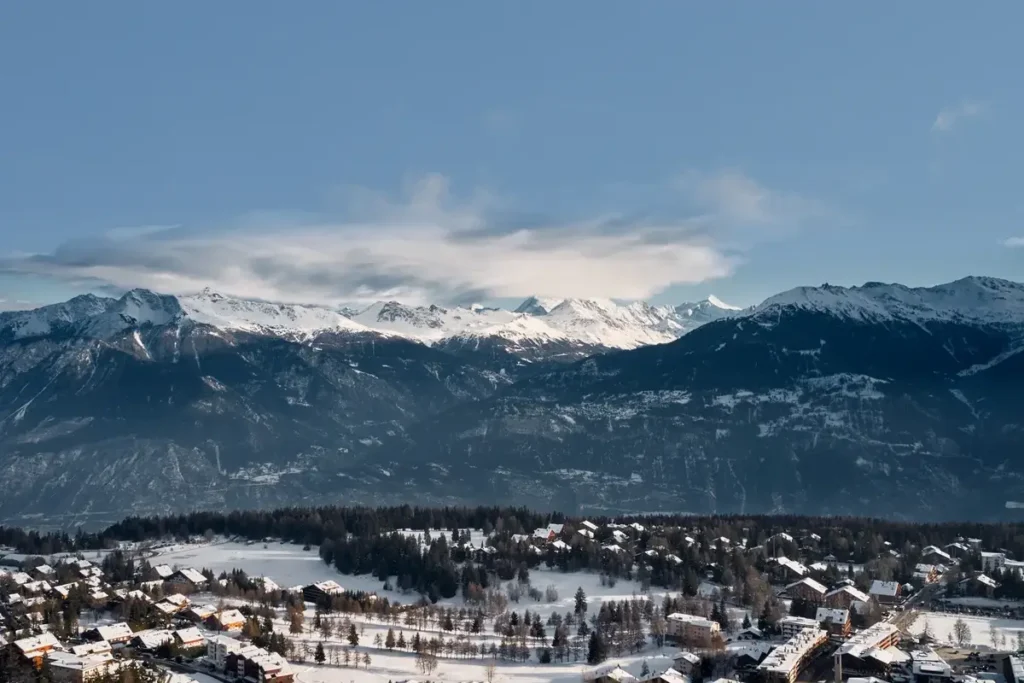 Snow-covered mountains rise beyond the buildings and wooded slopes of Crans-Montana.
