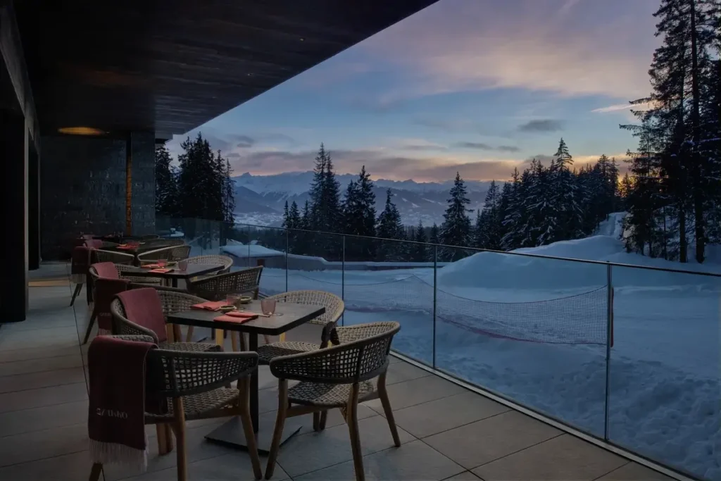 Dining tables sit beneath a dark canopy beside a glass railing overlooking snow and mountains at dusk.