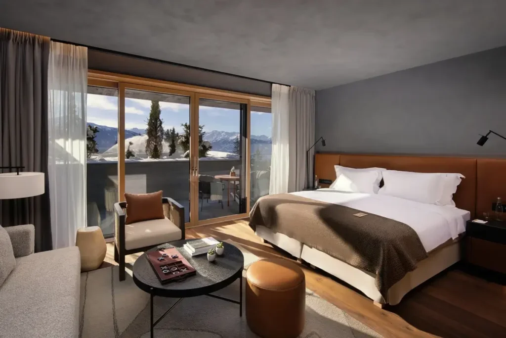A bed, lounge seating, and wood flooring face glass balcony doors with snowy mountain views.