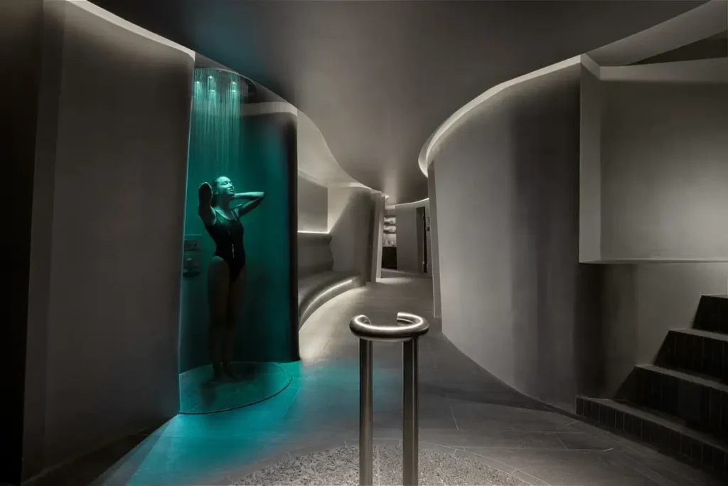 Curved gray walls surround a turquoise-lit shower alcove, a small basin, and steps in the spa wet area.
