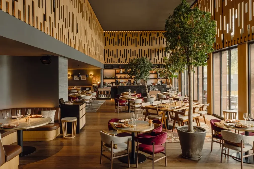Set dining tables, wood chairs with red cushions, and indoor trees beneath a patterned timber ceiling.