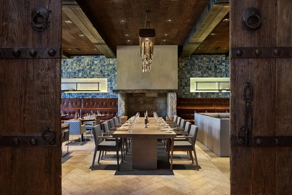 Large timber doors frame a long set dining table, upholstered chairs, blue patterned walls, and a fireplace.